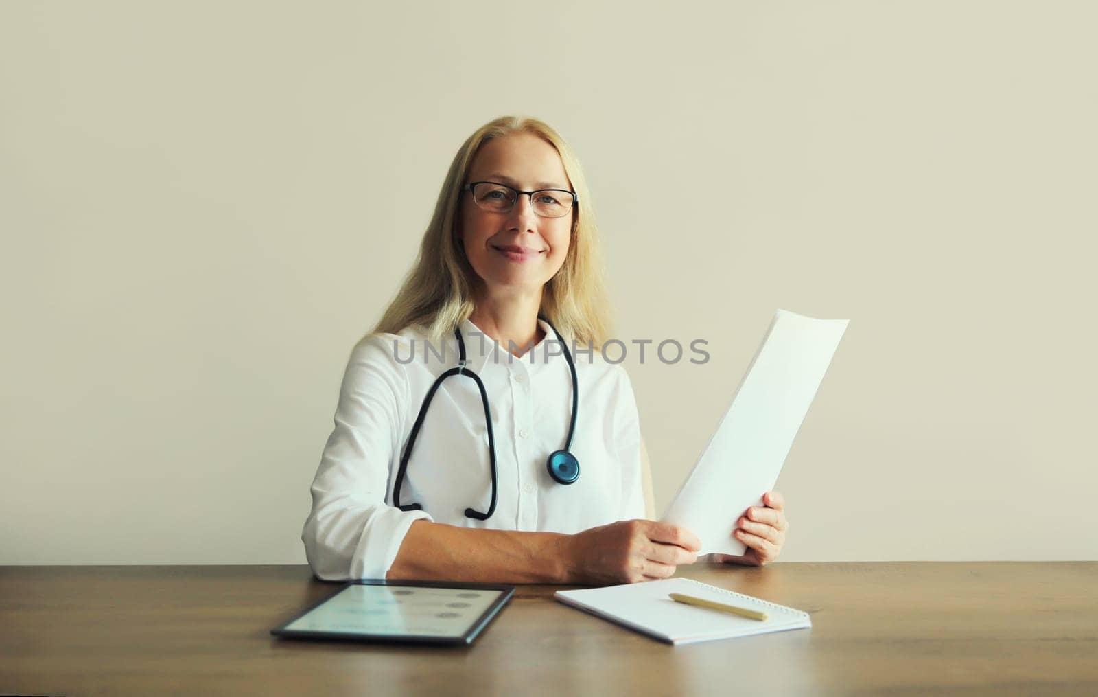 Caucasian middle-aged woman doctor with stethoscope, paper documents and digital tablet computer sitting at desk in clinic office