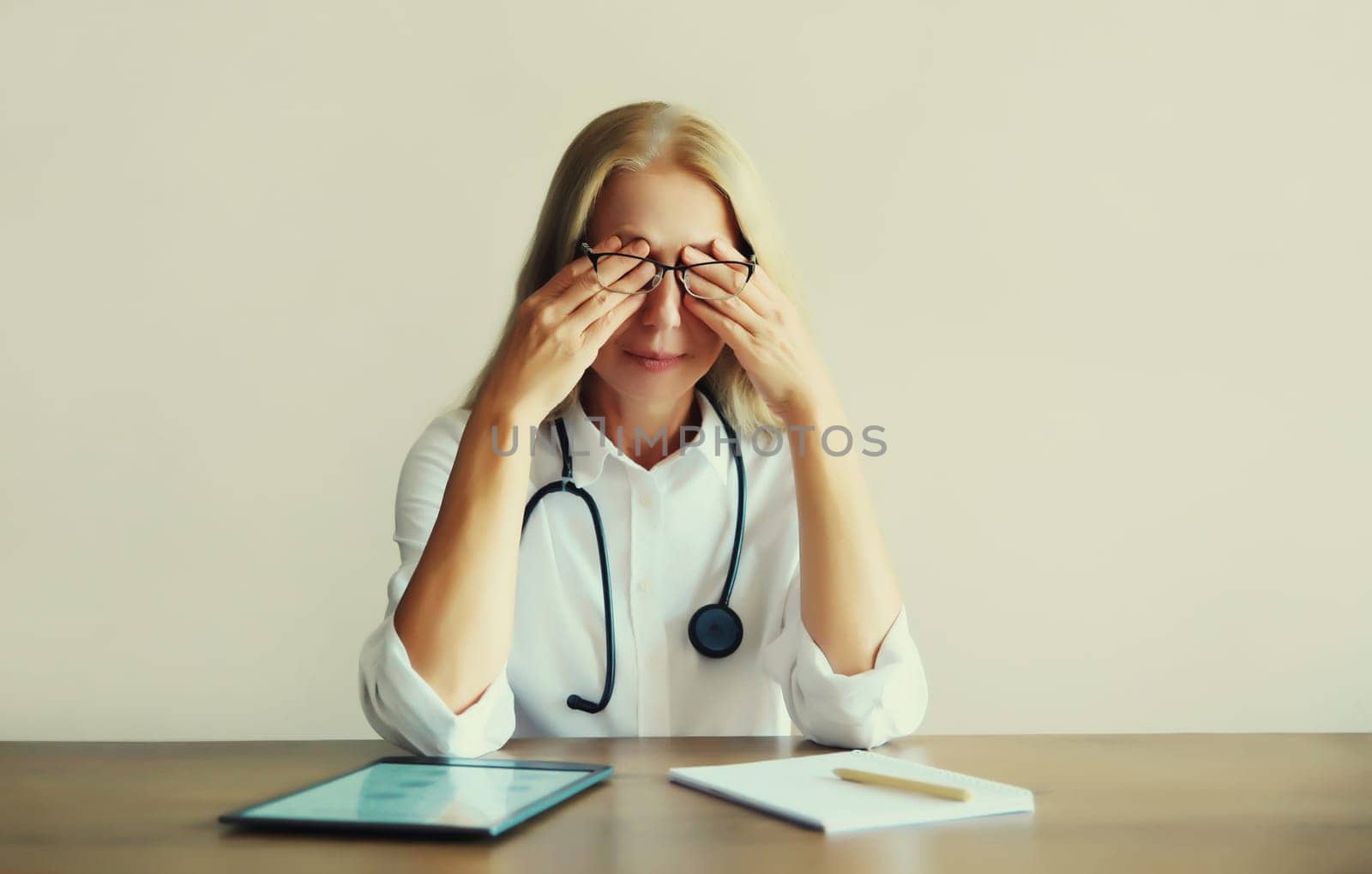Tired caucasian middle-aged woman doctor working with digital tablet and paper documents while sitting at desk in medical clinic office