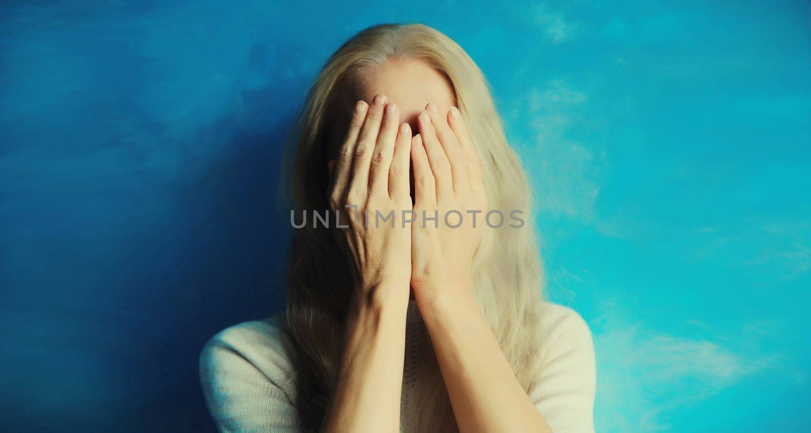 Upset sad crying middle-aged woman covering her eyes and face with her hands while experiencing mental or physical suffering sitting on couch in living room at home