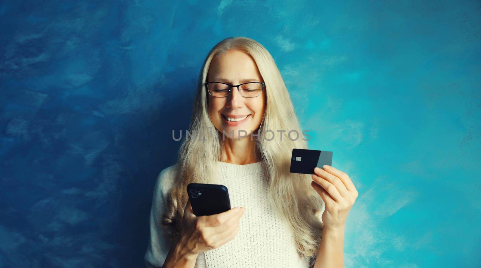Modern happy caucasian middle aged woman shopping online or ordering something looking at mobile phone holding plastic credit bank card in her hands