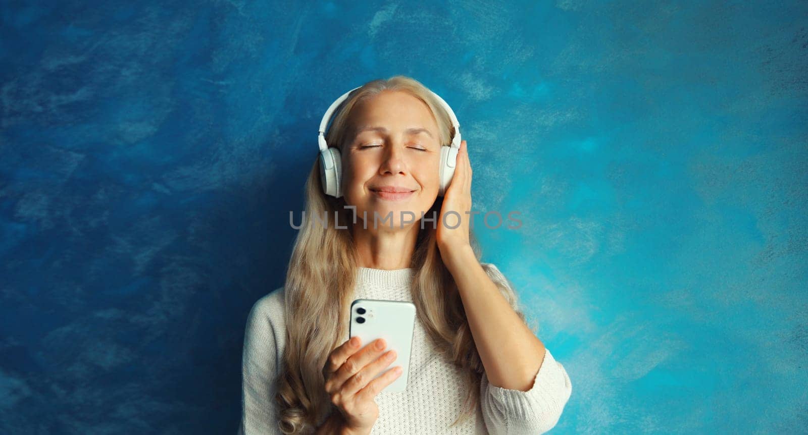 Portrait of happy smiling caucasian middle aged woman listening to music in headphones with smartphone on blue background