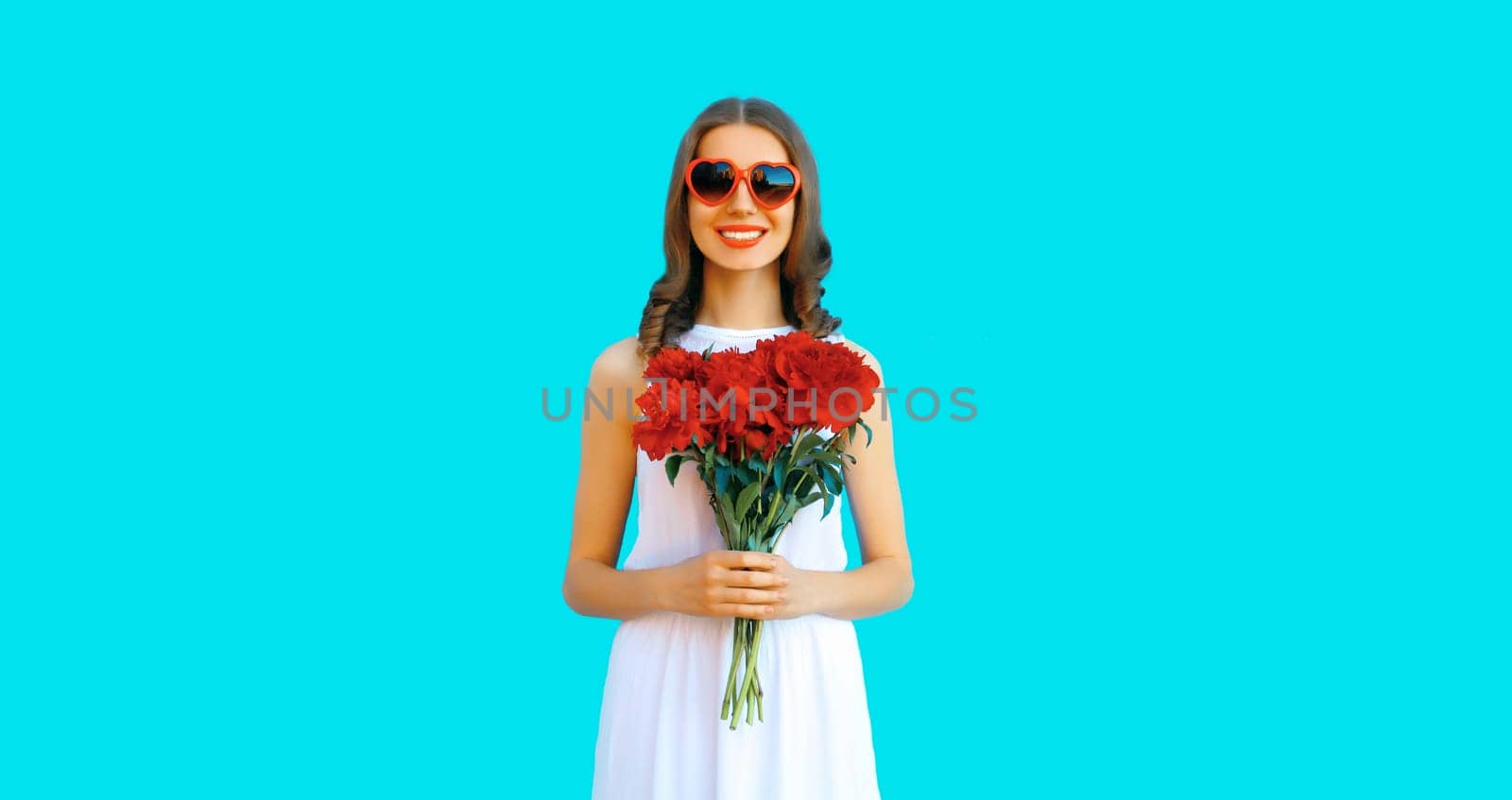Portrait of beautiful smiling woman with bouquet of red rose flowers in heart shaped sunglasses on blue background