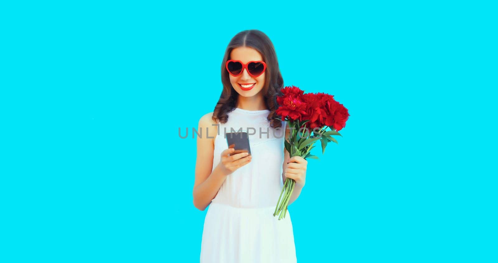 Portrait of beautiful smiling woman with phone and bouquet of red rose flowers in heart shaped sunglasses on blue background