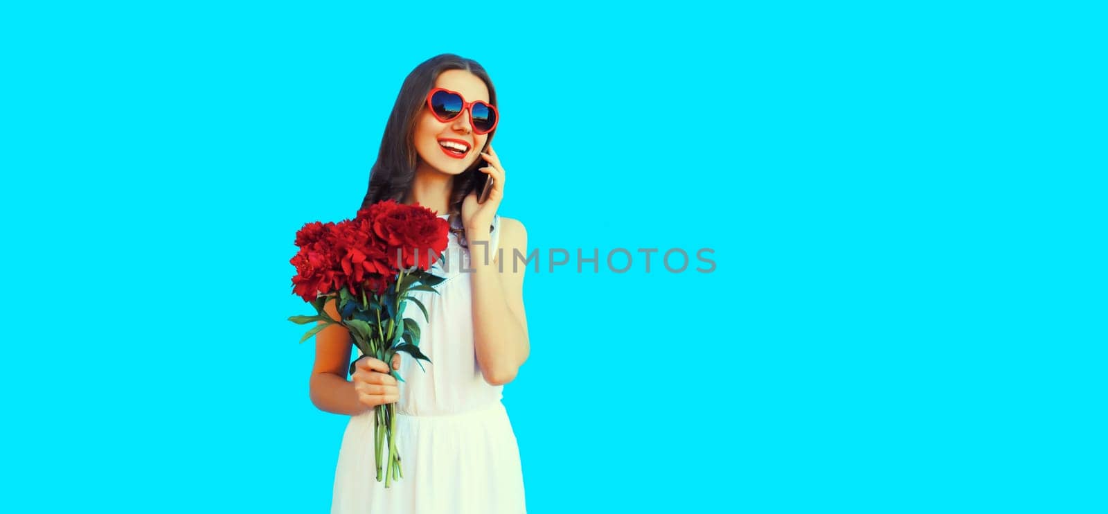Portrait of beautiful smiling woman calling on smartphone with bouquet of red rose flowers in heart shaped sunglasses on blue background
