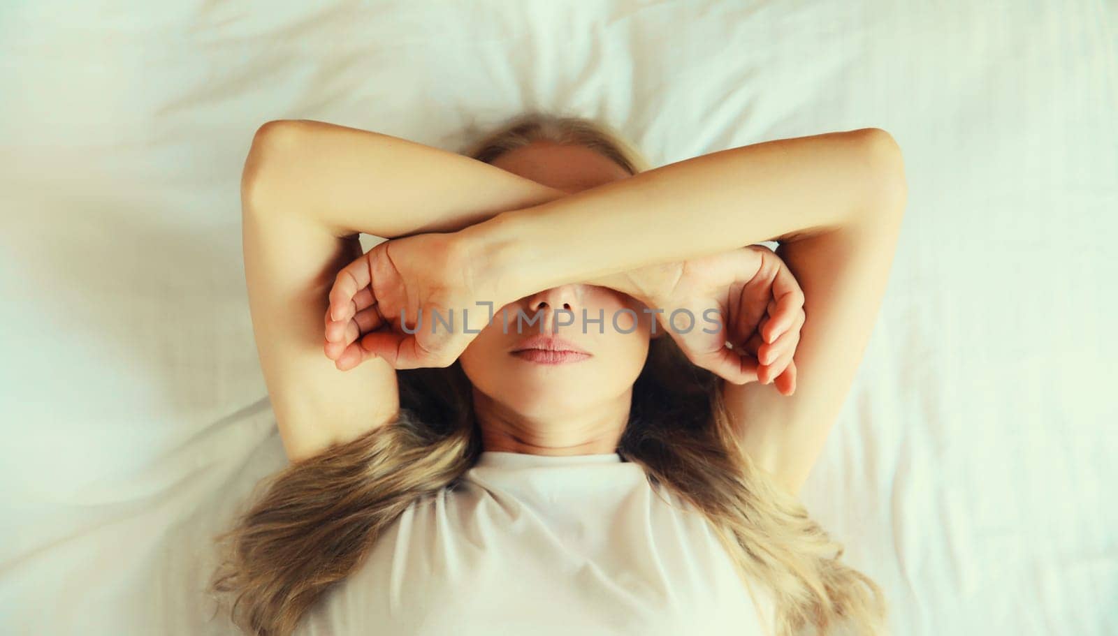 Tired sleepy woman wakes up covering her eyes with her hands lying on the bed on pillow in bedroom at home in the morning