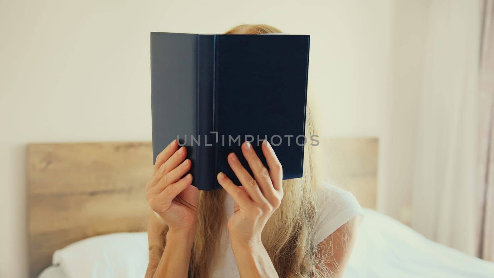 Young sad tired woman student covering her face with open book with blank cover while lying on the bed at home