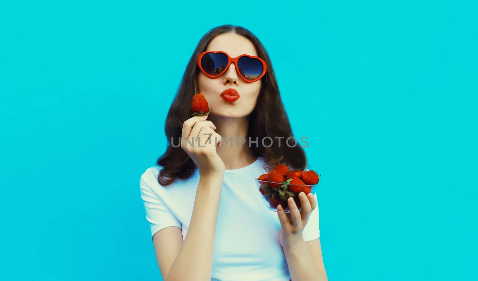 Portrait of beautiful caucasian young woman with handful of fresh strawberries blowing her lips wearing red heart shaped sunglasses on blue studio background