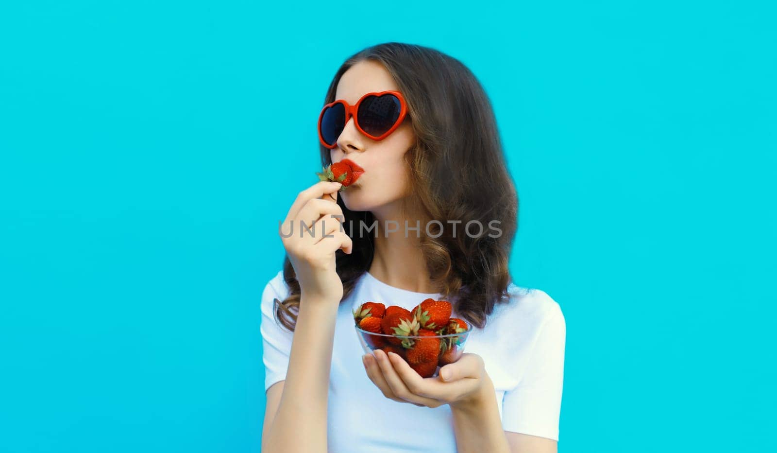 Portrait of beautiful caucasian young woman with handful of fresh strawberries blowing her lips wearing red heart shaped sunglasses on blue studio background