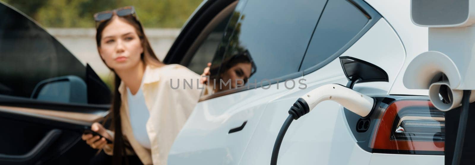 Young woman use smartphone to pay for electricity at public EV car charging station green city park. Modern environmental and sustainable urban lifestyle with EV vehicle. Panorama Expedient