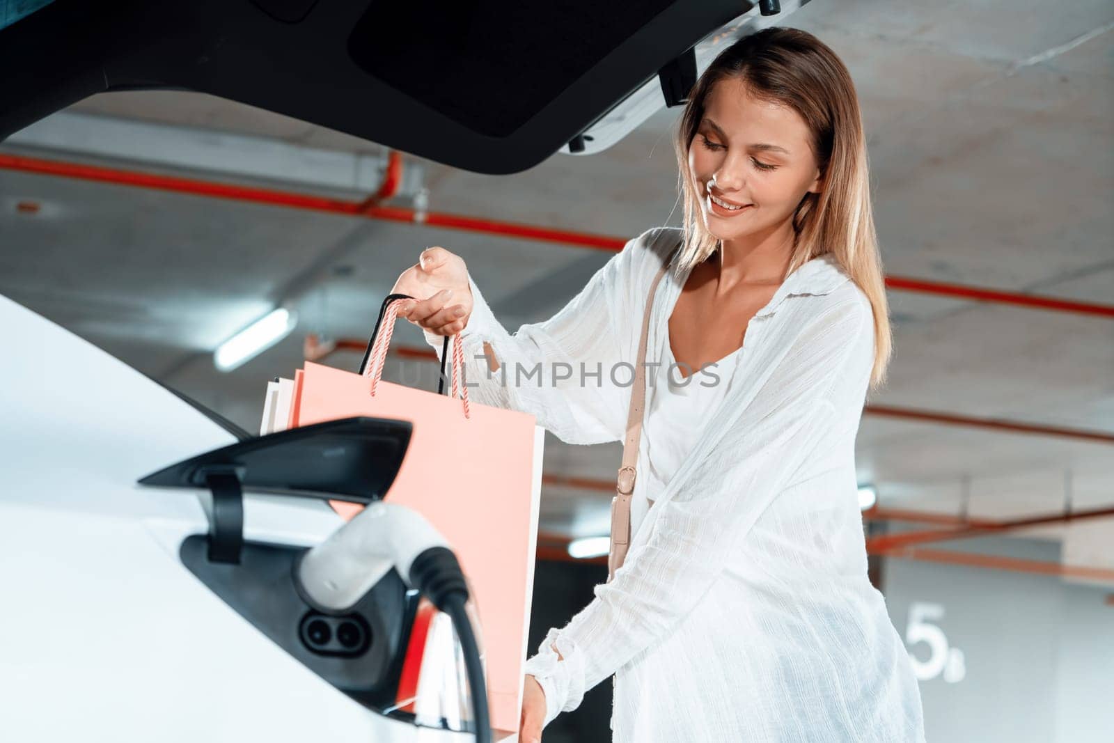 Young woman travel with EV electric car to shopping center parking lot charging in downtown city showing urban sustainability lifestyle by green clean rechargeable energy of electric vehicle innards