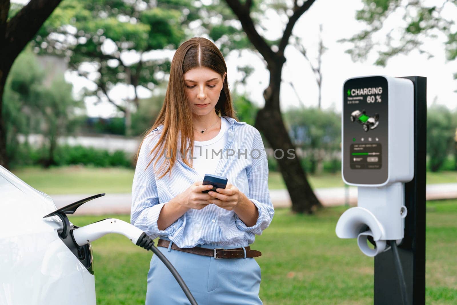 Young woman recharge EV electric vehicle battery from EV charging station and using smartphone online banking to pay for electricity in city park. Eco friendly vehicle travel with EV car. Exalt