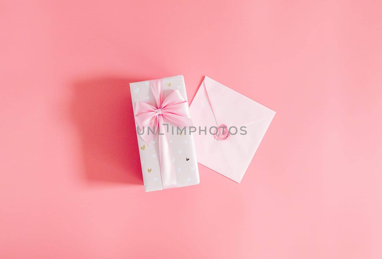 One beautiful gift box with a bow and a sealed envelope lie in the center on a pink background with copy space on the sides, flat lay close-up.