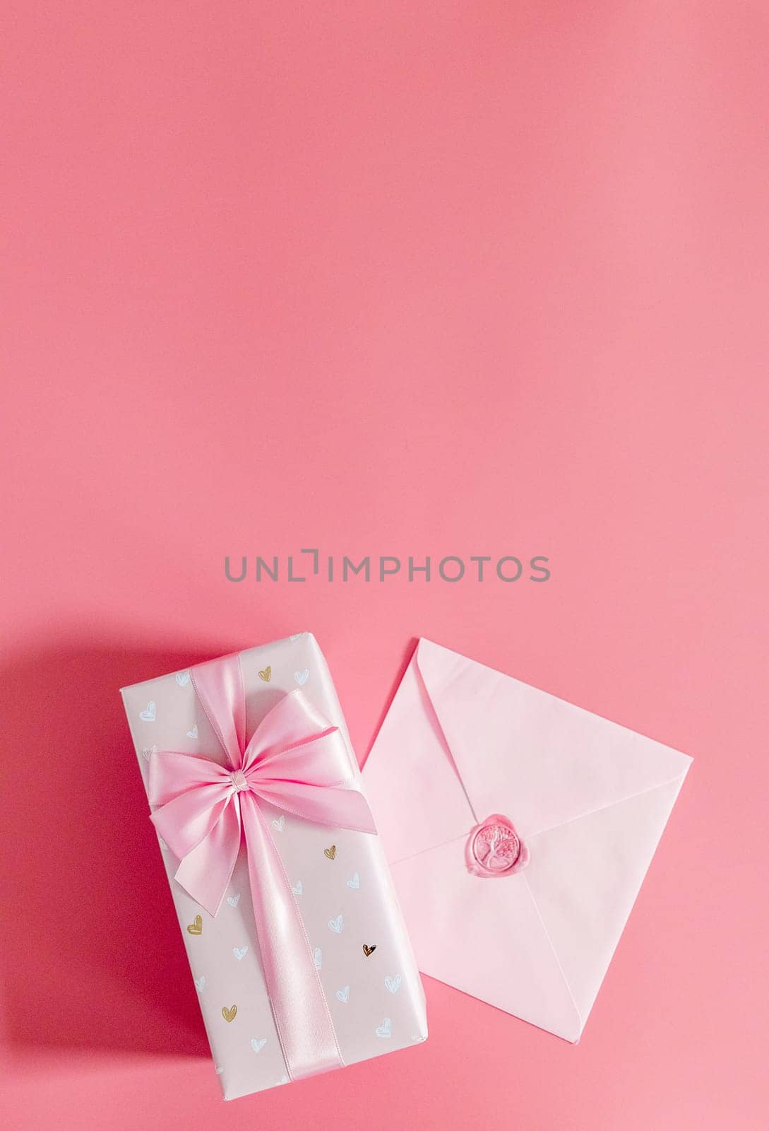 One beautiful gift box with a bow and a sealed envelope lie below on a pink background with copy space on top, flat lay close-up.