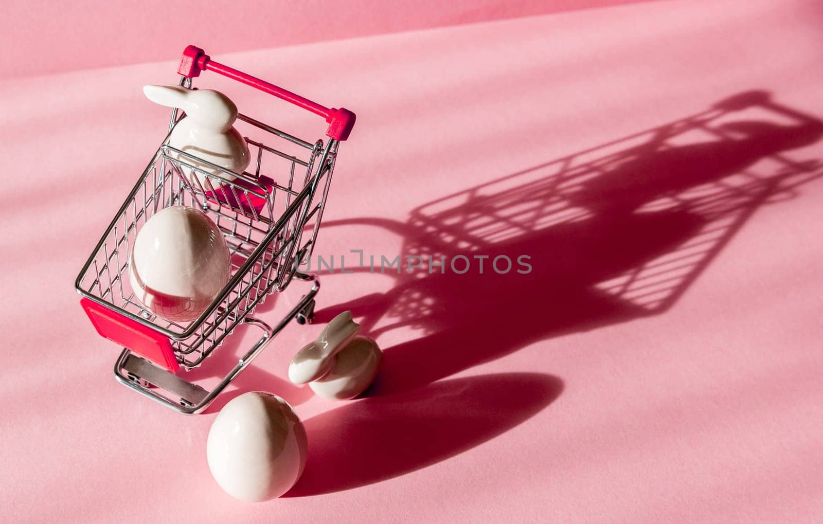 Porcelain figurines of Easter eggs and bunny with a mini shopping cart on the left on a pink background with shadows and copy space on the right, close-up top view.