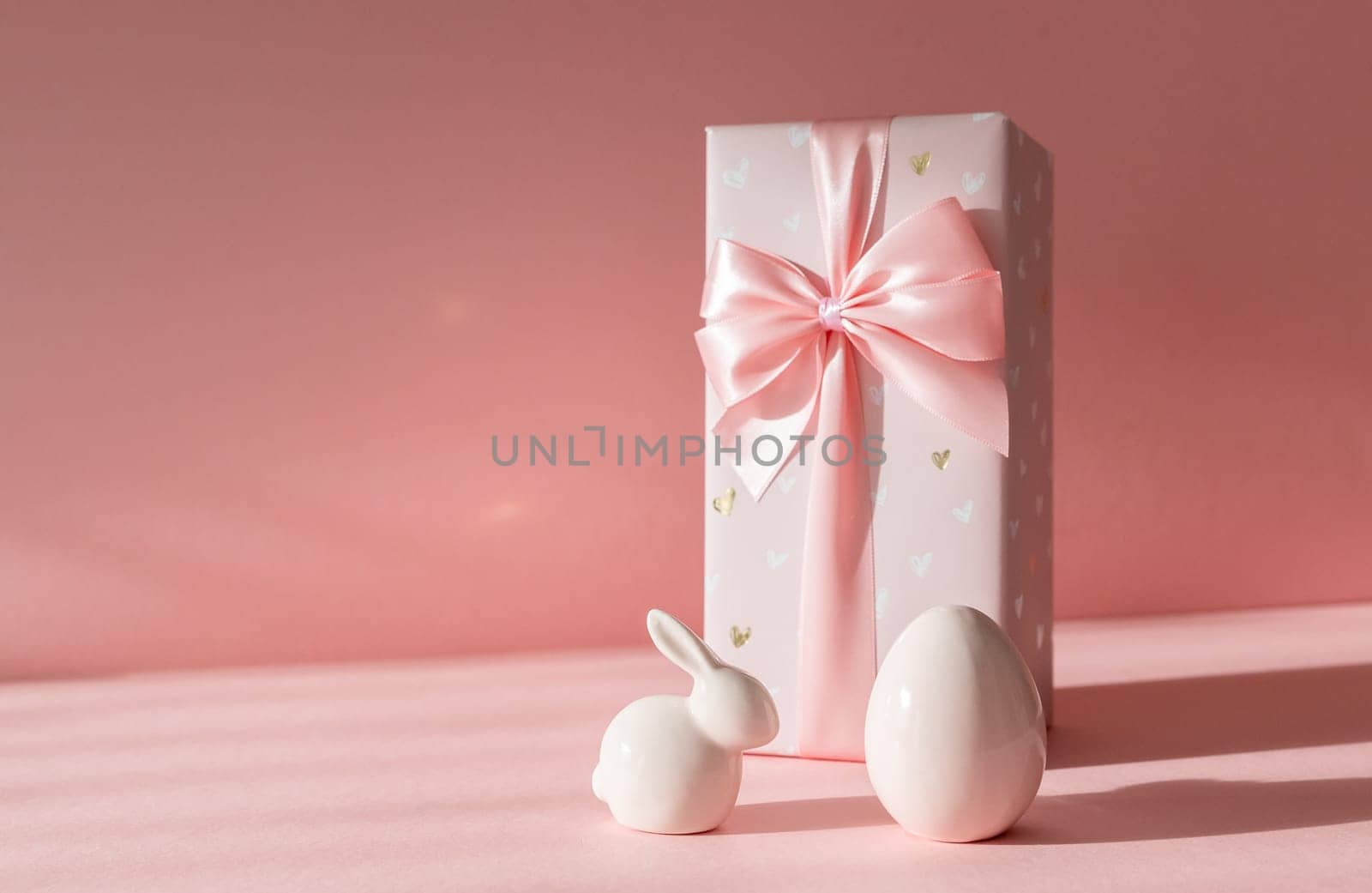 Porcelain figurines of Easter eggs and a big bunny gift box stand on the right on a pink background with shadows and copy space on the left, side view close-up.