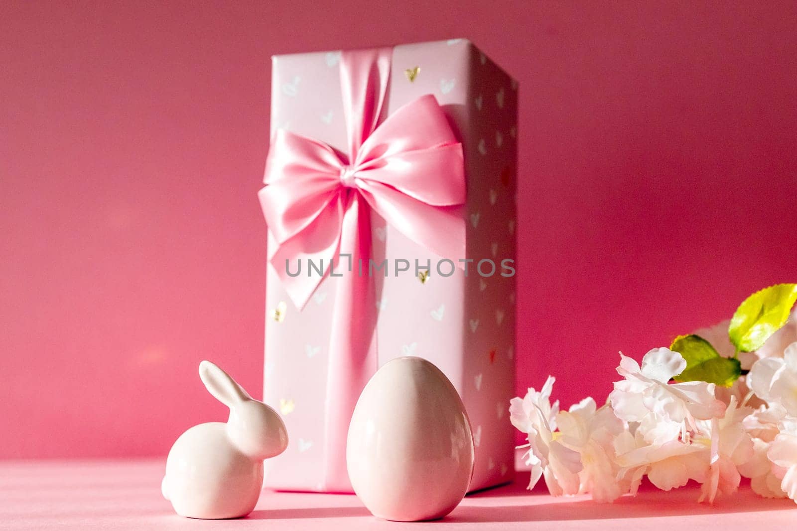 Porcelain figurines of Easter eggs, a bunny, a large gift box, and a branch of apple tree flowers stand on a pink background, close-up side view with selective focus.