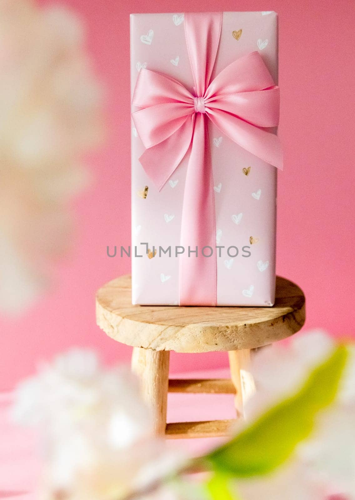 One large gift box with a bow stands on a small wooden stool on a pink background through the blurry branches of an apple tree, close-up side view.