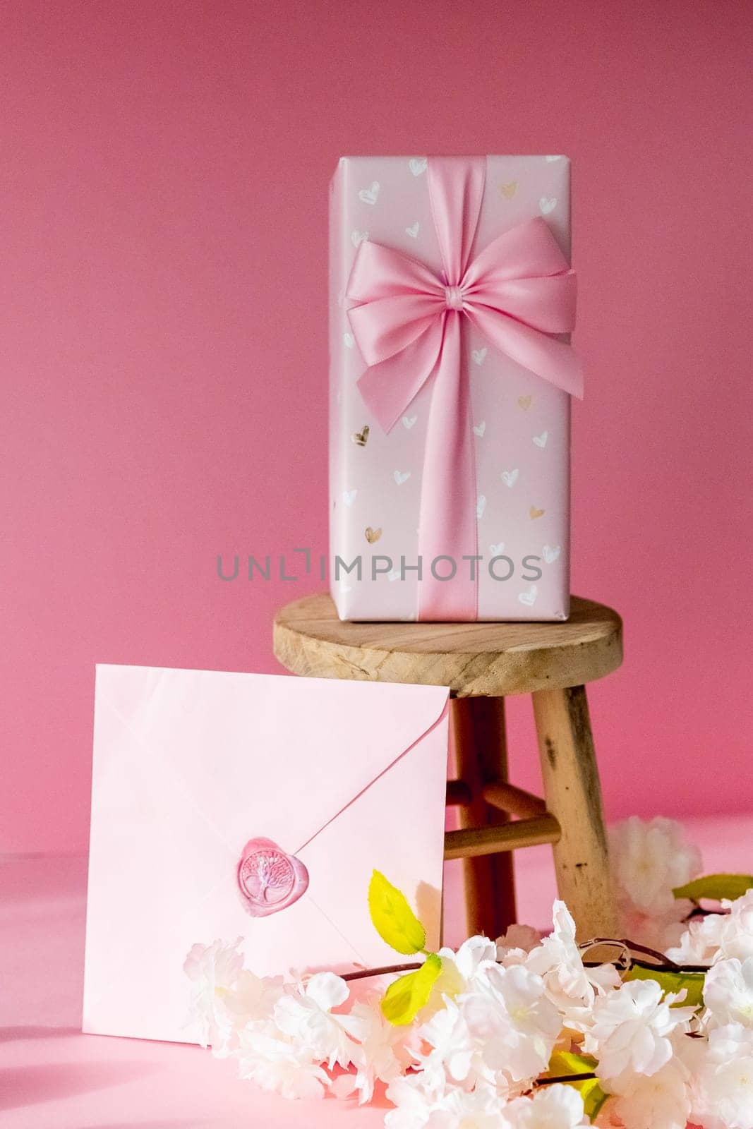 One sealed envelope, a large gift box with a bow stand on a small homemade decorative wooden stool on a pink background with a branch of apple tree blossom, side view close-up with selective focus.