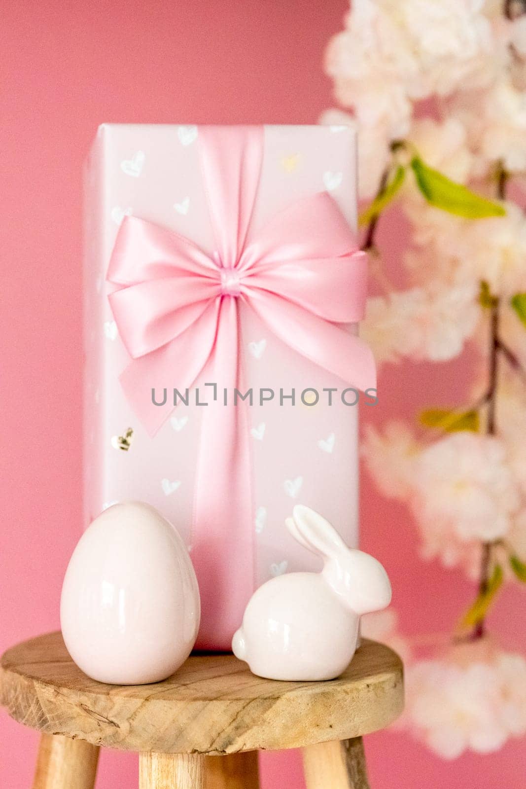 Porcelain Easter egg, bunny, large gift box with a bow stand on a small homemade decorative wooden stool on a pink background with a branch of apple tree blossom, side view close-up with selective focus.
