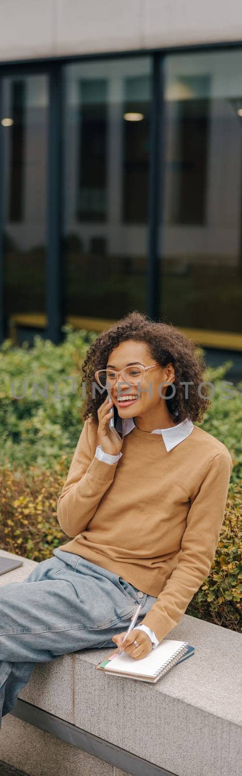 Smiling female freelancer talking phone and making notes while sitting on building background