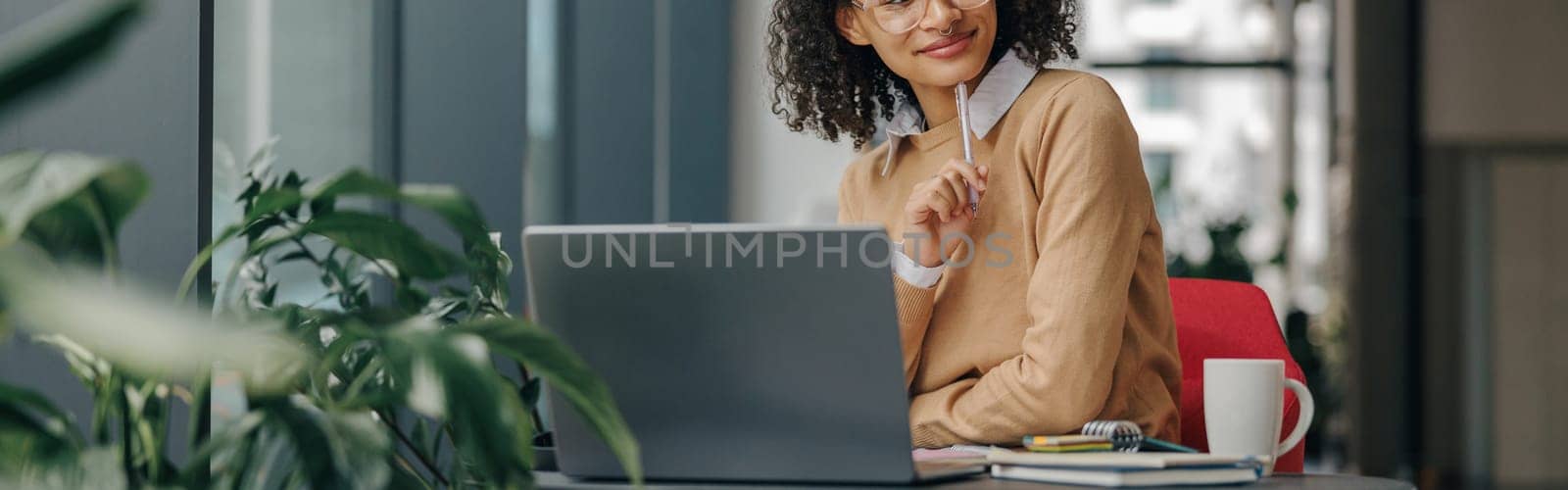 Pretty female manager in eyeglasses working on laptop while sitting in modern office near window