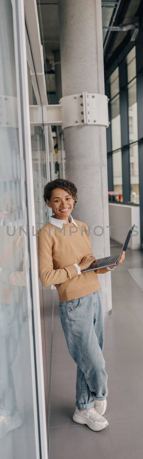 Smiling female manager using laptop while standing on office background and looks camera by Yaroslav_astakhov