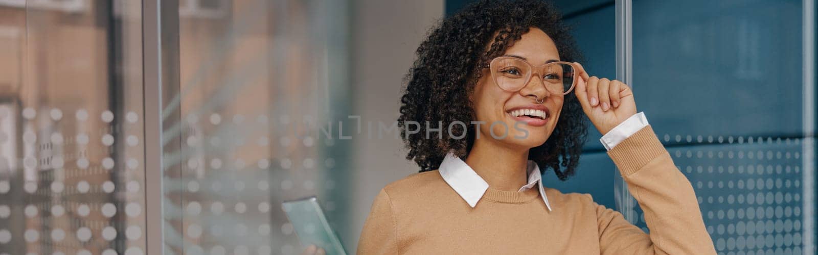 Smiling businesswoman holding phone while standing on modern office background and looks away by Yaroslav_astakhov