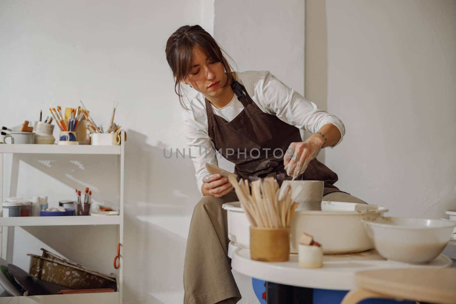 Focused female artisan in apron sitting on bench with pottery wheel and making clay pot by Yaroslav_astakhov