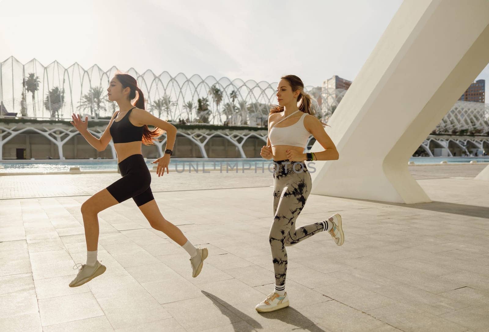 Two active women athlete running side by side along an outdoor track on modern buildings background