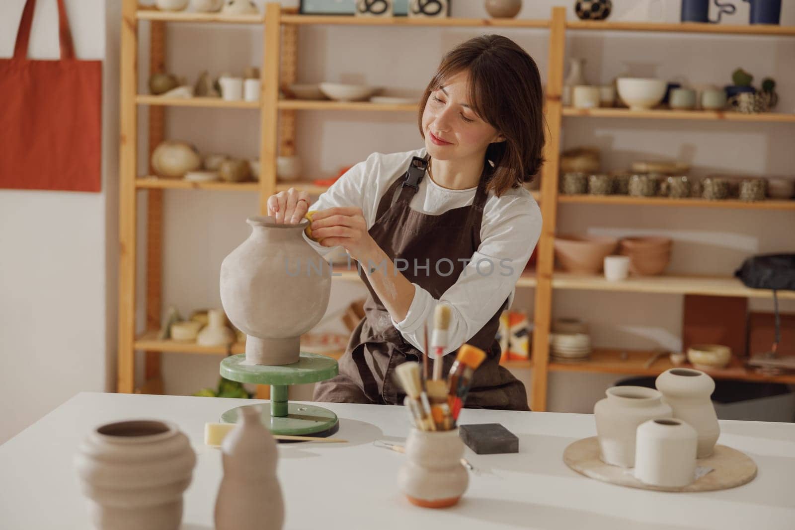 Smiling young female ceramist wearing apron work with unfired clay vase in pottery studio by Yaroslav_astakhov