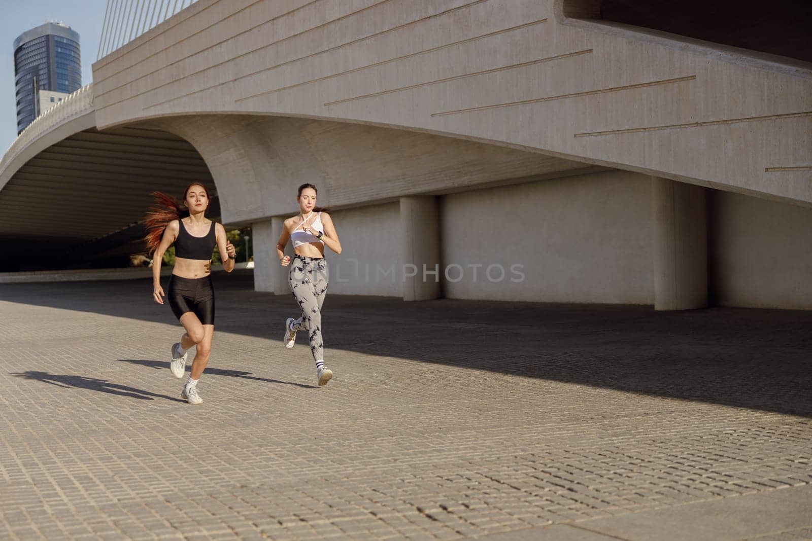 Two active women athlete running side by side along an outdoor track on modern building background by Yaroslav_astakhov