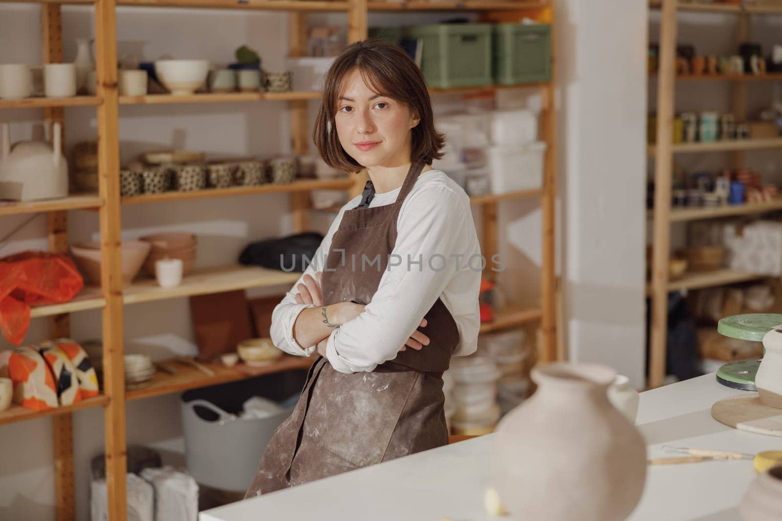 Portrait of young female potter in apron looking at camera while posing in workshop by Yaroslav_astakhov