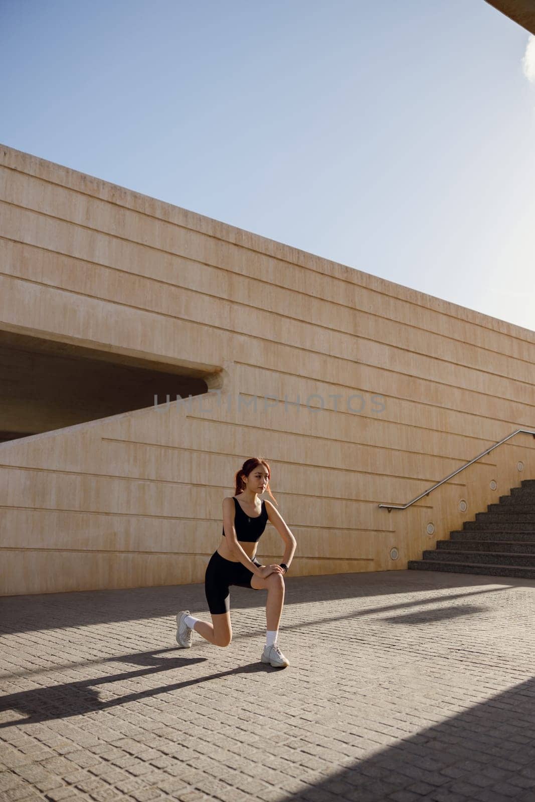 Young sportswoman in tracksuit stretches legs outside on modern building background by Yaroslav_astakhov