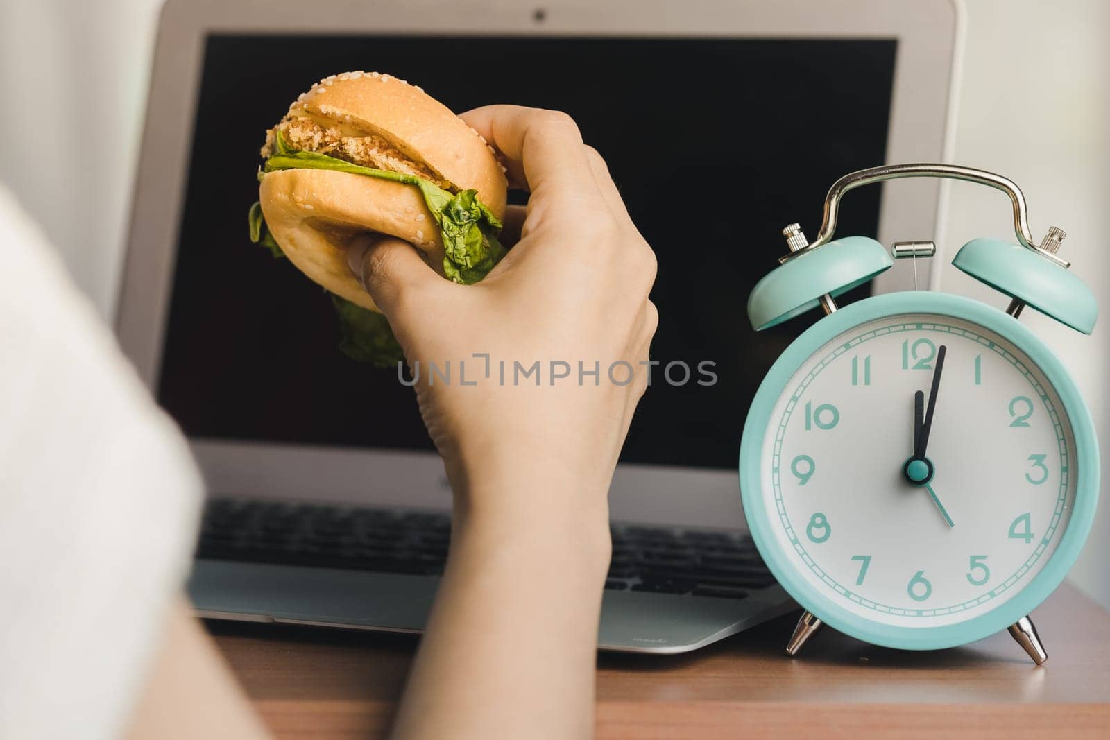 Person eating a hamburger while working, and there is an alarm clock displaying noon time on the work table by iamnoonmai