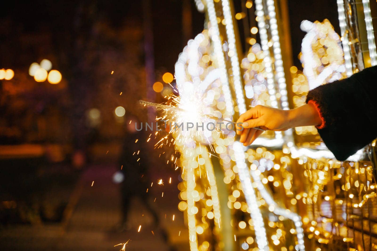 Woman holding sparkler night while celebrating Christmas outside. Dressed in a fur coat and a red headband. Blurred christmas decorations in the background. Selective focus.