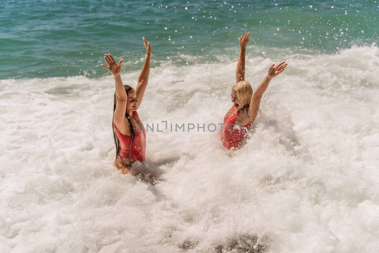 Women ocean play. Seaside, beach daytime, enjoying beach fun. Two women in red swimsuits enjoying themselves in the ocean waves and raising their hands up. by Matiunina