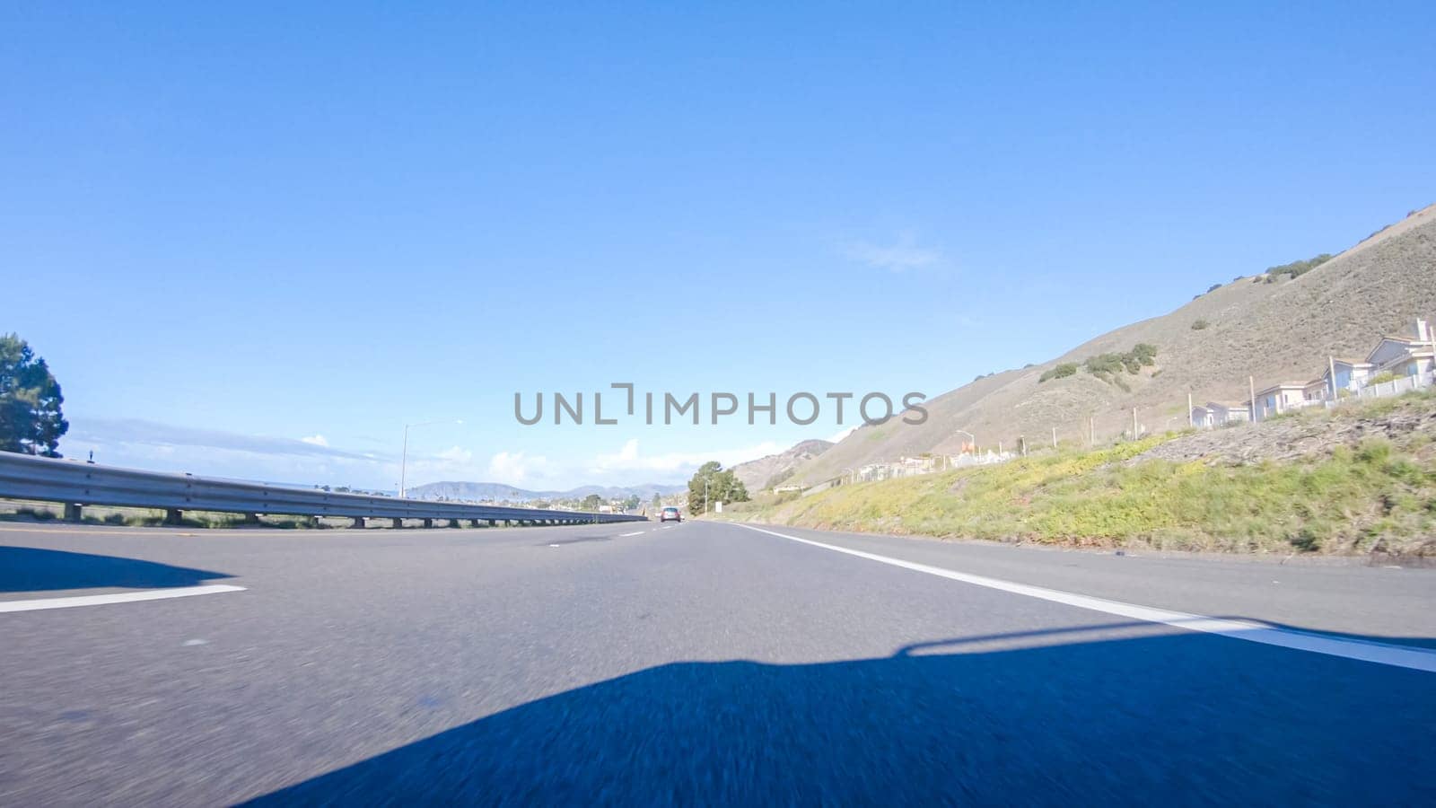 On a crisp winter day, a car cruises along the iconic Highway 1 near San Luis Obispo, California. The surrounding landscape is brownish and subdued, with rolling hills and patches of coastal vegetation flanking the winding road.