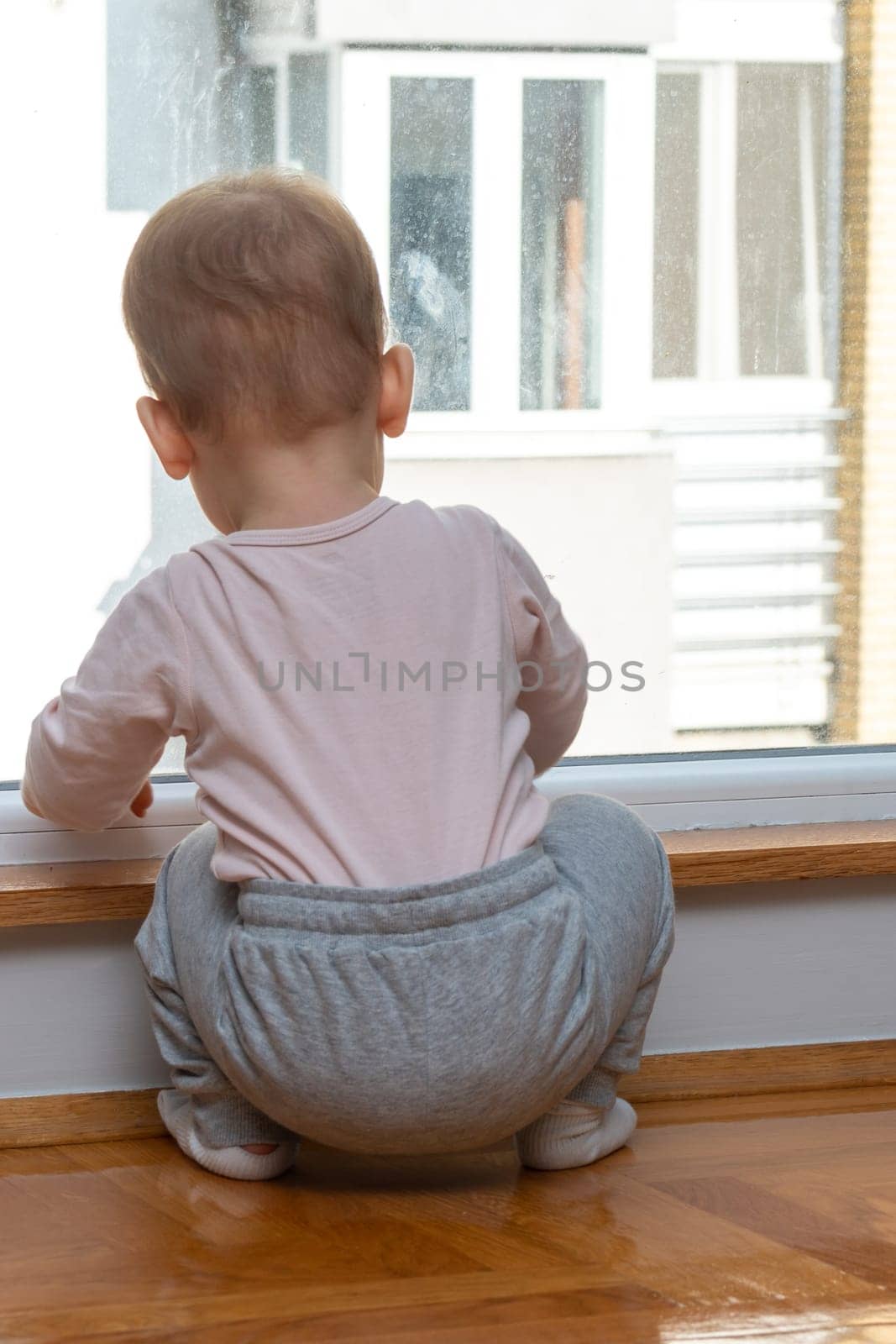 A toddler in a house looks out the window at a nearby building, dreaming of going outside for fun. Concept of a kid wanting to leave home for a walk