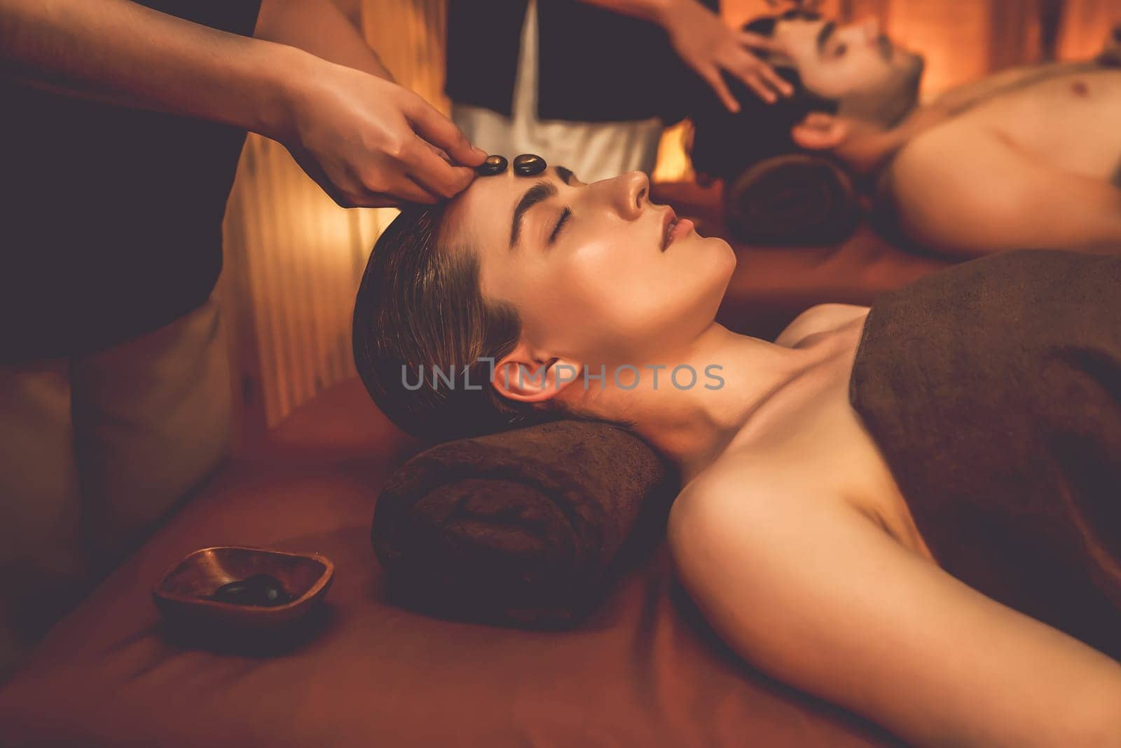 Caucasian couple enjoying relaxing anti-stress head massage with hot stone and pampering facial beauty skin recreation leisure in warm candle light ambient salon spa in resort or hotel. Quiescent