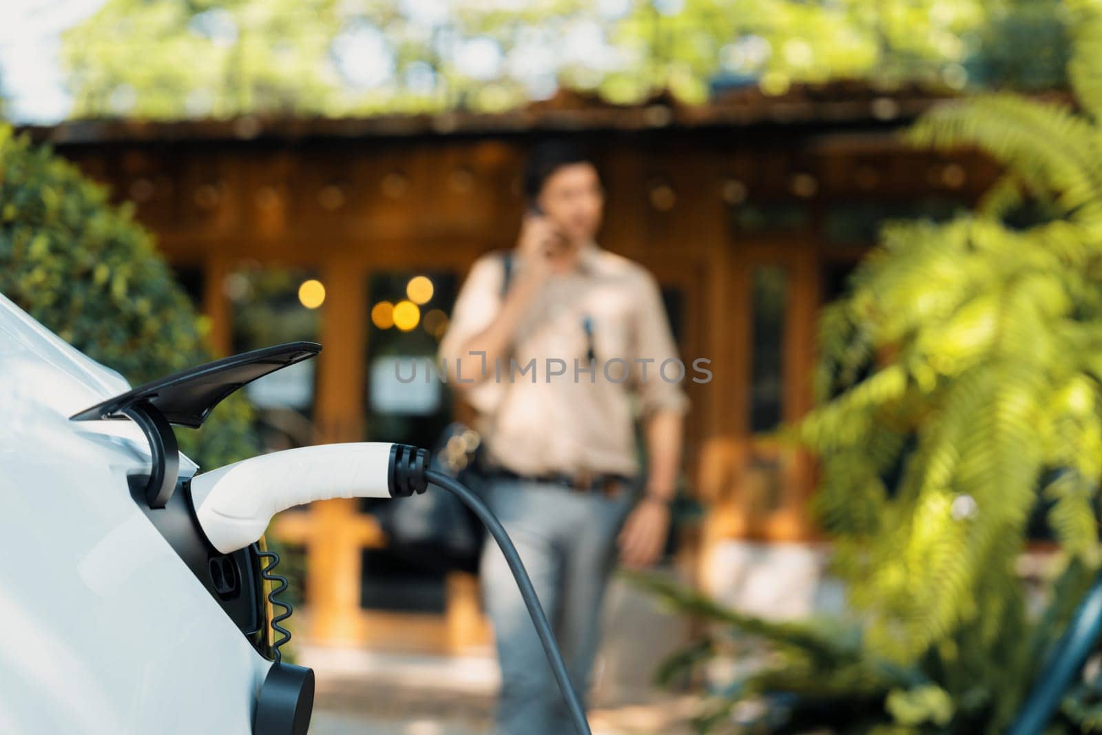 Focused EV electric car recharging at outdoor coffee cafe in springtime garden with blur background of eco friendly man, green city sustainability and environmental friendly EV vehicle. Expedient