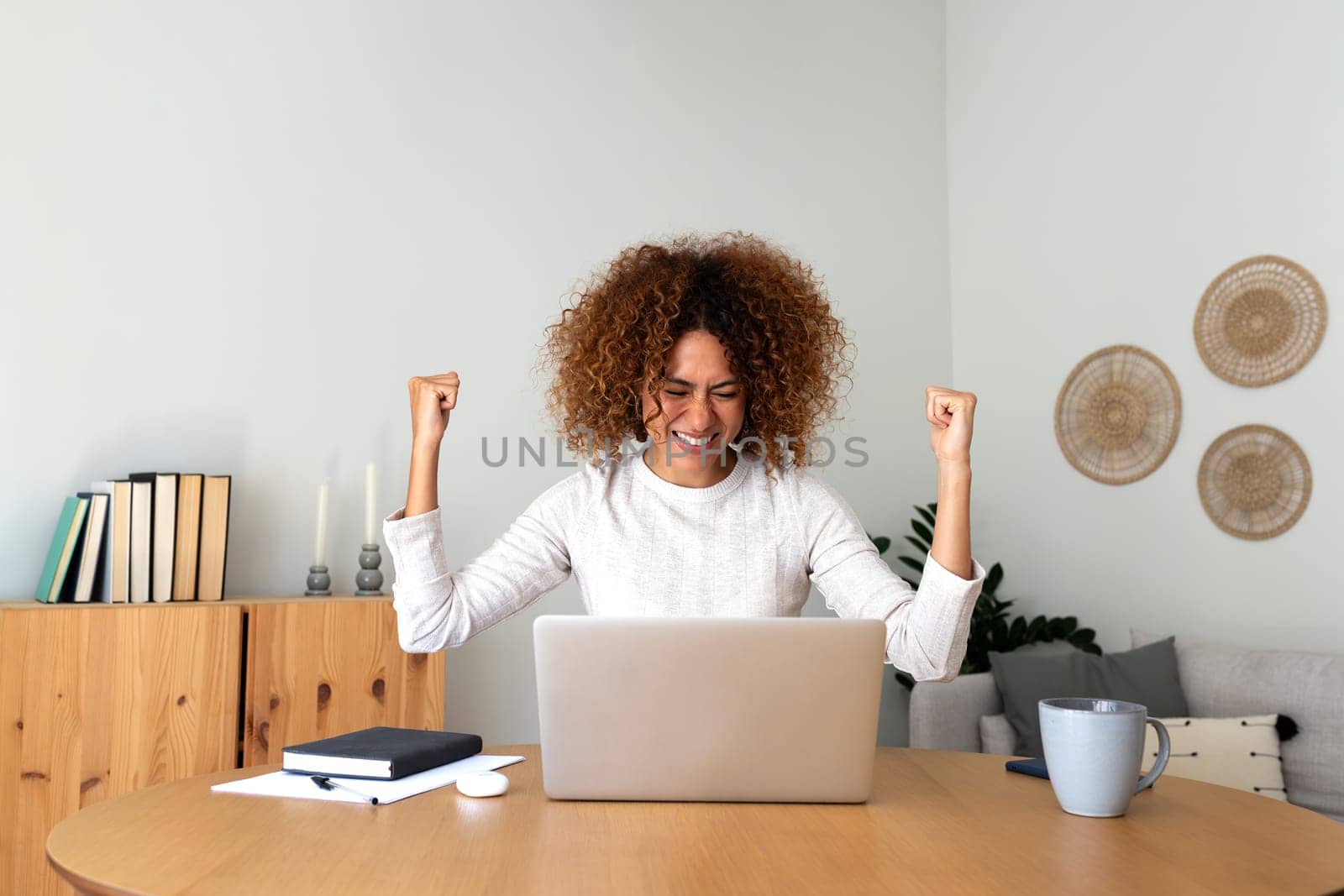 Happy young multiracial Hispanic female celebrating success with arms raised up in front of laptop at home office. by Hoverstock