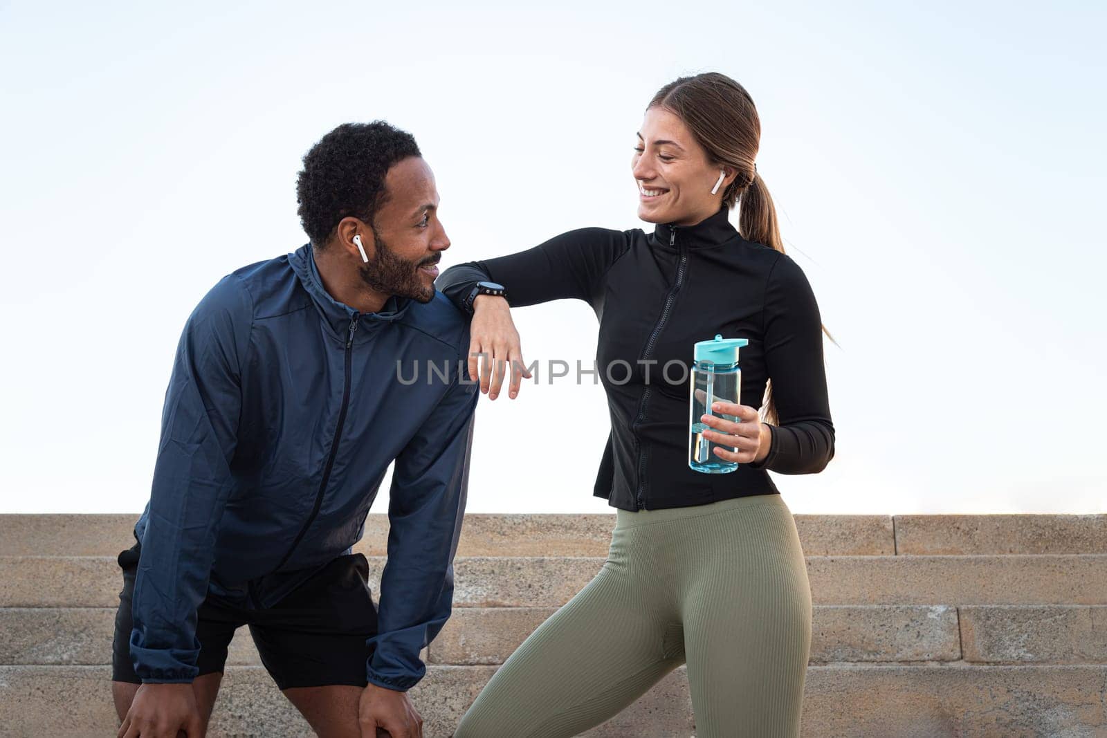 Young happy multiracial couple taking a break after running and working out together outdoors. Female holding water bottle. Fitness and sport concept.
