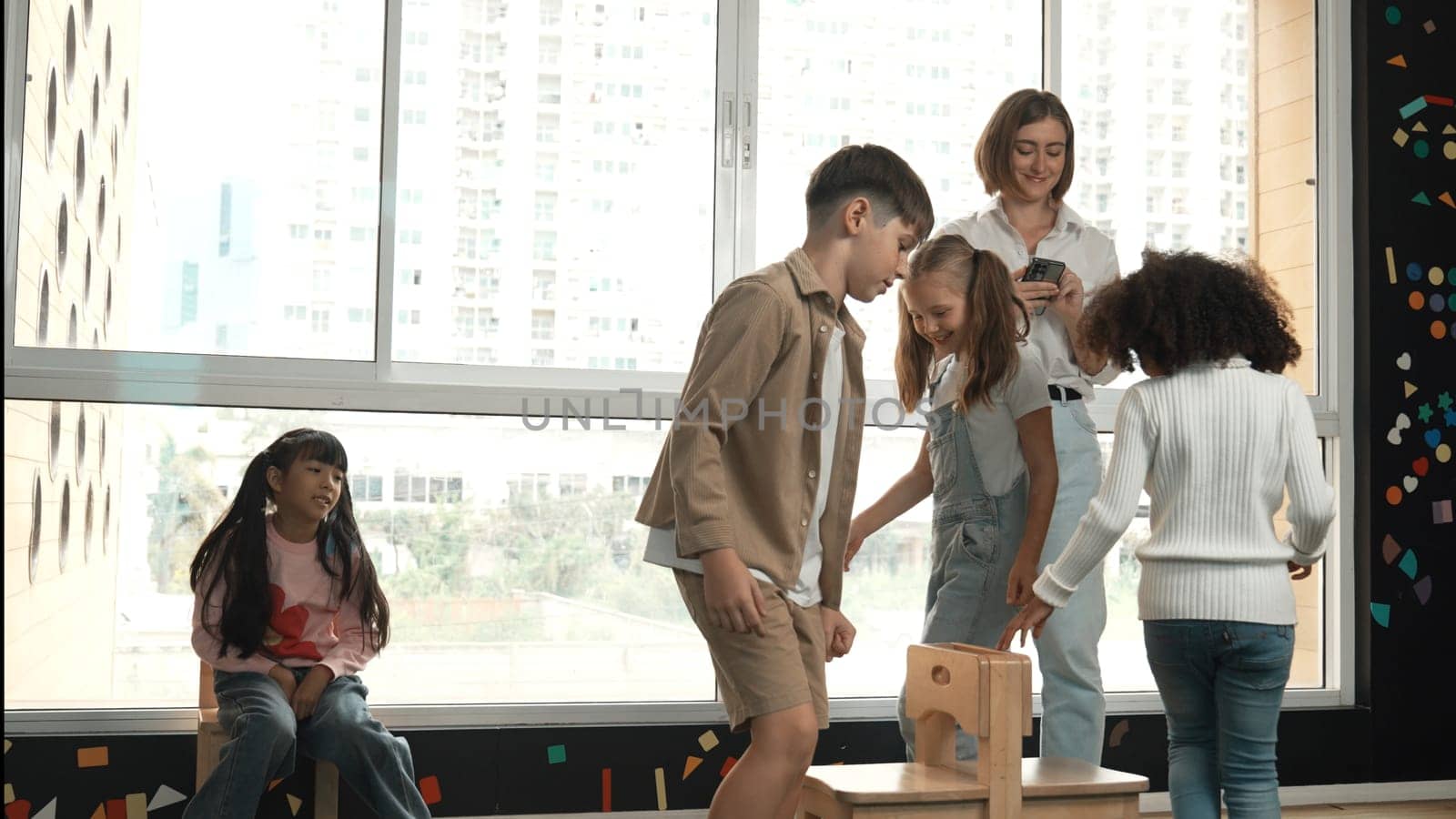 Diverse children playing musical chairs games while young smart beautiful teacher turn off music. Multicultural students running and sitting on chair in creative activity. Music lesson. Erudition.