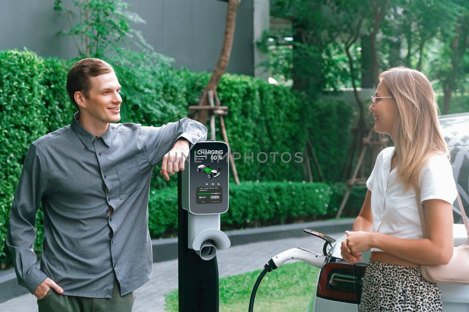 Young couple travel with EV electric car charging in green sustainable city outdoor garden in summer shows urban sustainability lifestyle by green clean rechargeable energy of electric vehicle innards