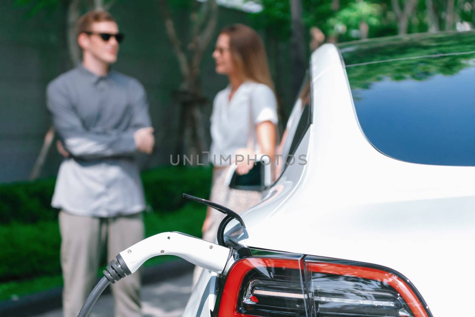 Young couple travel with EV electric car charging in green sustainable city outdoor garden in summer shows urban sustainability lifestyle by green clean rechargeable energy of electric vehicle innards