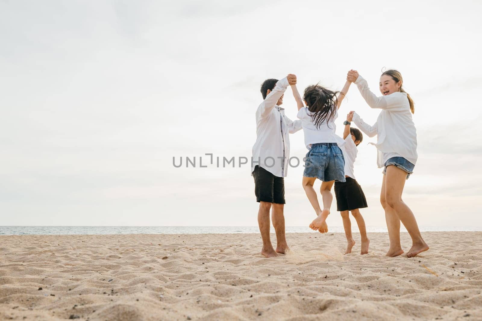 A burst of joy on beach as an Asian family father and mother holding children leaps into air. beautiful ocean provides serene setting for this delightful portrayal of family togetherness. by Sorapop