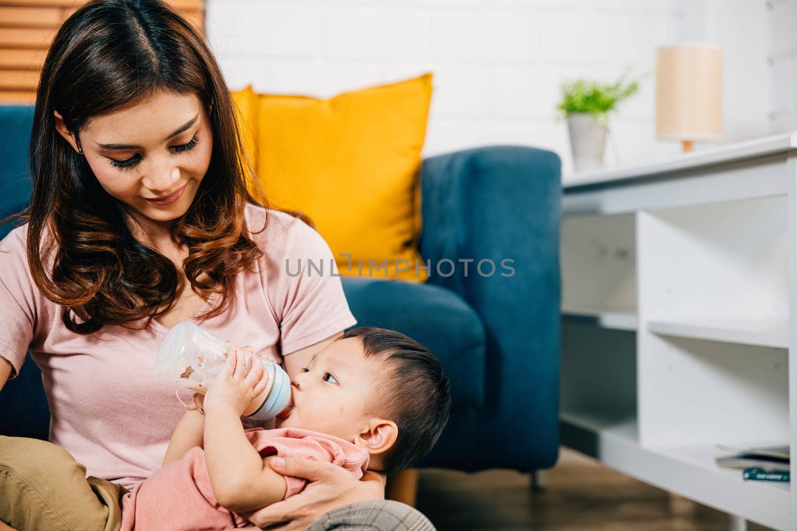 A heartwarming family portrait comes to life in the living room at home as a smiling mother holds her cute Asian baby who is feeding from a baby bottle radiating joy and togetherness in every glance.