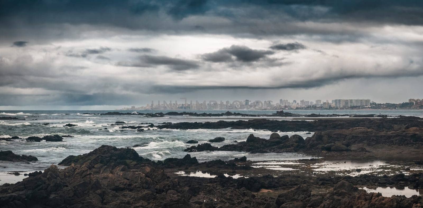 Dramatic clouds hang above a rocky coastline with a far-off urban skyline of Salvador de Bahia