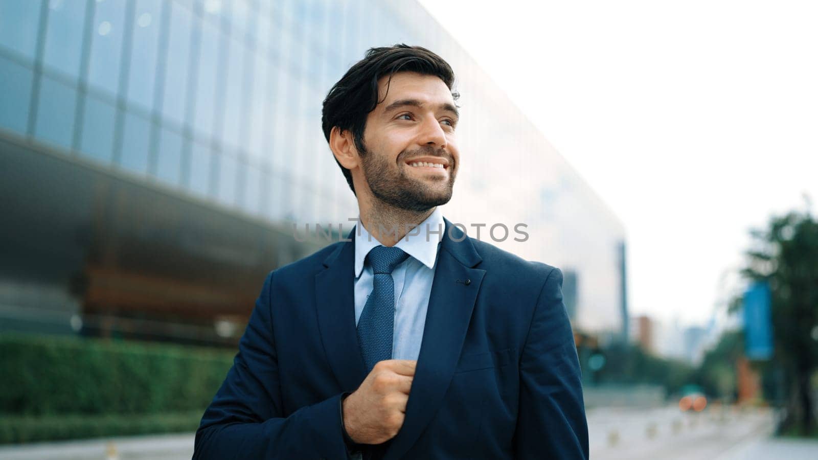 Portrait of professional smiling business man standing at camera at city view. Skilled project manager standing with cool pose with blurred background. Handsome investor pose at camera. Exultant.