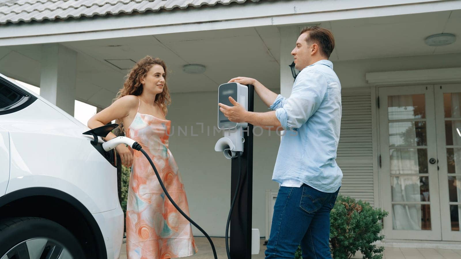 Happy and lovely couple with eco-friendly conscious recharging electric vehicle from EV charging station. EV car technology utilized as alternative transportation for future sustainability. Synchronos